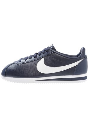 Dunkelblauer Nike-Sneaker aus Leder mit weißem Swoosh-Logo, weiß-blau gestreifter Sohle und passenden dunkelblauen Schnürsenkeln, seitlich gezeigt.
