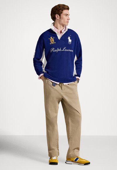 Mladý muž stojící se založenýma rukama v kapsách, oblečený v modré rugby košili Ralph Lauren, béžových kalhotách, růžové košili a žlutých teniskách na bílém pozadí.