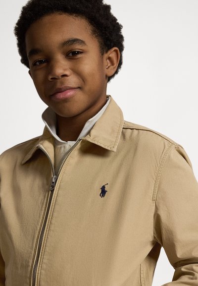 Beige glidelåsjakke med oppbrettet krage, med en liten marineblå logo på venstre bryst og en glatt, vevd tekstur.