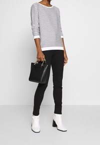 Maglione a righe grigio e bianco, pantaloni slim-fit neri, stivaletti bianchi alla caviglia, tiene una piccola borsa nera con dettagli in oro.