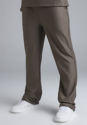 Bruine geribbelde broek gecombineerd met witte vetersneakers, gedragen door een persoon die tegen een effen achtergrond staat.