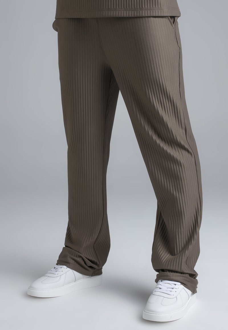 Pruunid soonikkoes püksid koos valgete paeltega tossudega inimesel, kes seisab ühevärvilise tausta ees.