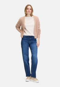 Lichtroze gebreide cardigan over een grijs gestreept shirt, gecombineerd met blauwe spijkerbroeken en beige sneakers. De cardigan heeft korte mouwen.