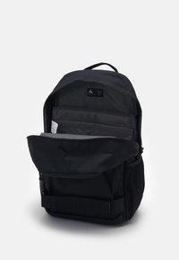 Lo zaino nero presenta un compartimento principale con zip, una tasca laterale in rete e spallacci regolabili. L'interno include diverse sezioni imbottite.