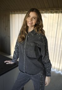Tumši pelēks džinsu krekliņš ar priekšējo kabatu, rotāts ar rhinestones pie piedurknēm. Nēsāts kopā ar atbilstošām tumši pelēkām biksēm.