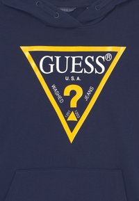 Sudadera azul marino con el logotipo de Guess en forma de triángulo contorneado en amarillo, que presenta un gran signo de interrogación y el texto "GUESS U.S.A. Washed Jeans."