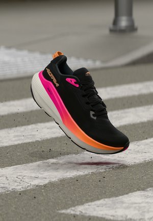 Zapatilla de correr negra con suela degradada en rosa y naranja, flotando sobre un paso de peatones en una ciudad sobre asfalto gris.