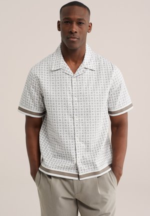 Homme portant une chemise blanche à manches courtes avec un motif et des bordures gris et blanc, associée à un pantalon gris clair, debout les mains dans les poches.