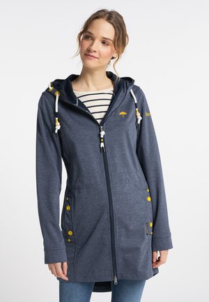 Frau trägt einen langen marineblauen Kapuzenpullover mit Reißverschluss und gelben Knöpfen und darunter ein gestreiftes Shirt, steht vor einem schlichten weißen Hintergrund.