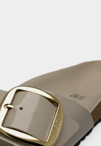 Μπεζ σανδάλι με γυαλιστερό επάνω υλικό, μεγάλο λουρί με χρυσή αγκράφα και καφέ υφή στο πάτο. Μινιμαλιστικός σχεδιασμός με καμπυλωτές άκρες.