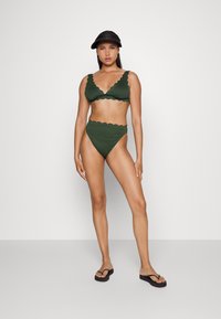 Conjunto de bikini verde com bordas em forma de ondas; top tipo bralette e parte de baixo de cintura alta. Modelo combinado com chinelos pretos e um boné.