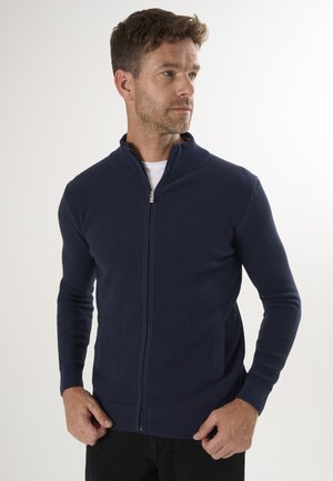 Pull à fermeture éclair navy avec texture côtelée, col haut et poches latérales. Design ajusté avec des manches longues et des finitions tricotées aux poignets et à l'ourlet.