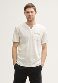 Witte short-sleeve shirt met een henley-kraag en een zak op de linkerborst. Gemaakt van een zachte, gestructureerde stof. Gepaard met donkere broeken.