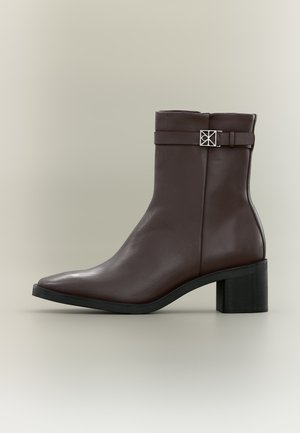 Dunkelbrauner Leder-Stiefelette mit Blockabsatz, spitzer Schuhspitze und dekorativem Riemen mit silberner quadratischer Schnalle auf neutralem Hintergrund.