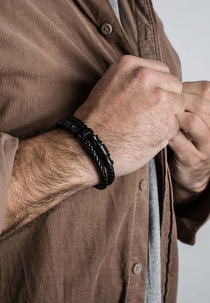 Man's clasped hands showing black braided leather bracelet with cylindrical clasp, brown corduroy shirt, and gray undershirt.
