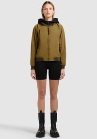 Junge Frau steht, trägt eine olivgrüne Jacke mit Reißverschluss und schwarzer Kapuze, schwarze Radlerhosen und schwarze klobige Stiefel, vor einem schlichten hellen Hintergrund.