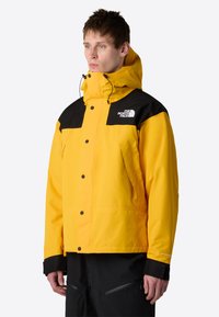 Žlutá vodotěsná bunda s černými akcenty, zipovými kapsami a nastavitelnou kapucí, s výrazným logem na hrudi. Hladký povrch a robustní knoflíky.