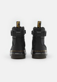 Bottines noires avec une tige en cuir lisse, des panneaux latéraux en mesh et une semelle chunky translucide. Elles comportent une languette jaune et des accents de boucle.