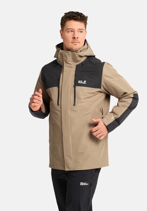 Man draagt een beige en zwarte Jack Wolfskin hooded jas met ritszakken, poseert met handen dicht bij borst en taille.