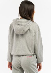 Grau beschnittenes Sweatshirt mit Kapuze, auf dem Rücken mit einem schwarzen Logo versehen. Kombiniert mit passenden grauen Hosen, die von pinken Buchstaben akzentuiert werden.