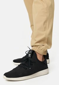 Zapatillas de deporte negras de punto con un acabado texturizado, suela de goma blanca y diseño de cordones. Combinadas con pantalones joggers de color beige claro y puños.