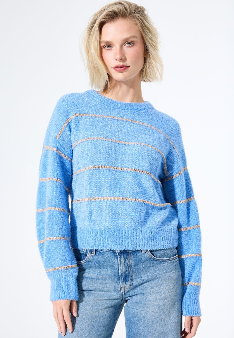 Helles blaues Strickpullover aus strukturiertem Stoff, mit horizontalen goldgestreiften Akzenten und gerippten Bündchen, kombiniert mit hellblauen Jeans.
