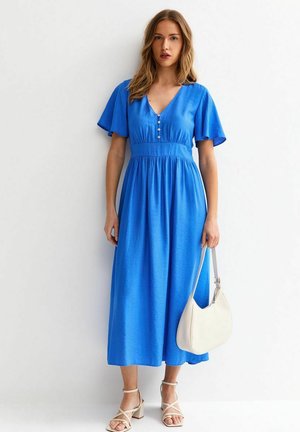 Kobieta stojąca oparta o białą ścianę, ubrana w niebieską sukienkę midi z guzikami i falbaniastymi rękawami, trzymająca białą torebkę i nosząca beżowe sandały.