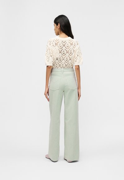 Γυναίκα με μακριά σκούρα μαλλιά που φοράει λευκό πλεκτό πουλόβερ με κοντά μανίκια και ανοιχτό πράσινο παντελόνι ψηλόμεσο και φαρδύ, με την πλάτη γυρισμένη.