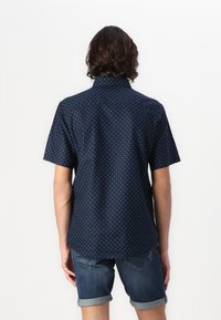 Chemise à manches courtes, couleur marine, avec de petits motifs en triangles blancs. Elle possède un col, une patte de boutonnage et un ourlet arrondi. Portée avec un short en denim.
