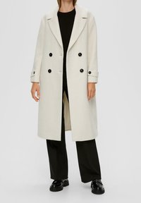 Kofta i ullblandning med lång krämfärgad design, utrustad med kavajkrage, dubbelknäppt stängning med svarta knappar och sidofickor, bärs över svart topp och byxor.