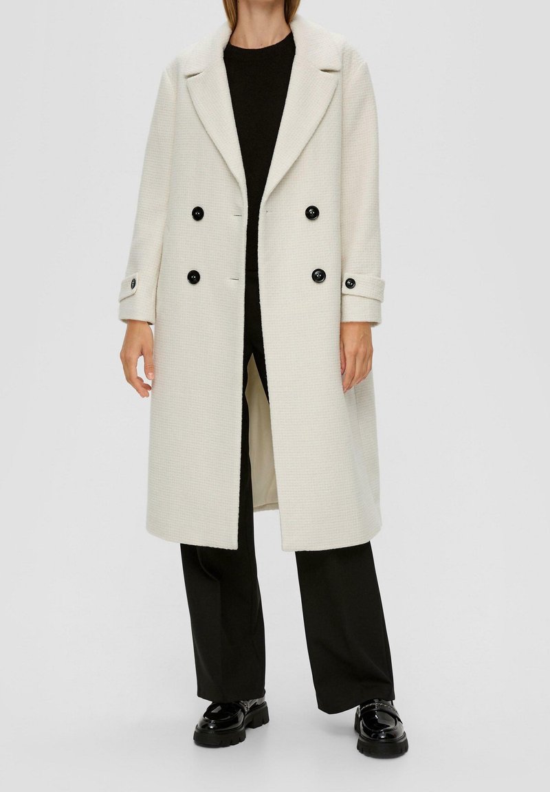 Kofta i ullblandning med lång krämfärgad design, utrustad med kavajkrage, dubbelknäppt stängning med svarta knappar och sidofickor, bärs över svart topp och byxor.