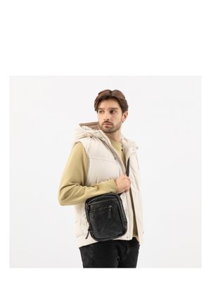Mann trägt eine beige ärmellose Weste und ein grünes Langarmhemd und hält eine schwarze Leder-Umhängetasche vor weißem Hintergrund.