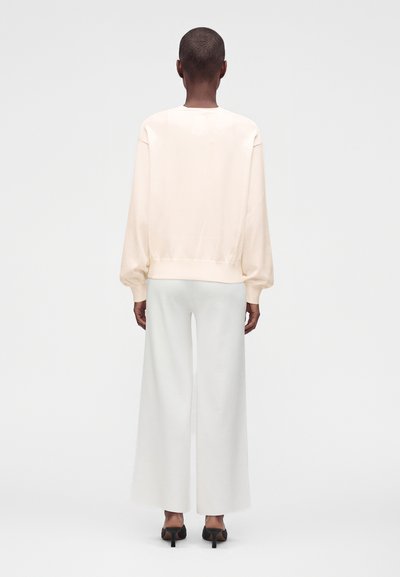 Personne aux cheveux courts portant un haut crème à manches longues et un pantalon blanc large, debout de dos sur un fond clair uni.