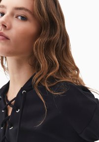 Černá mikina s kapucí a šněrováním vpředu se stříbrnými očky. Hladká tkanina s volným střihem a měkkou texturou, která zdůrazňuje přirozené vlny ve vlasech.