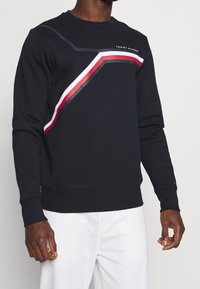 Navy crew neck sweatshirt made of cotton, featuring diagonal red and white stripes, and logo on the chest; ribbed cuffs and hem.