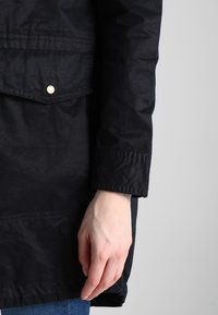 Černá nepromokavá parka s přední kapsou, zlatým drukem, dlouhými rukávy a hladkou texturou, s volným střihem vhodným pro vrstvení.