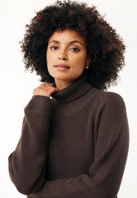 Maglione a collo alto in maglia marrone con texture a coste, spalle scese e vestibilità comoda. La modella tiene delicatamente il collo, evidenziando il capo.