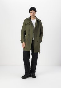 Abrigo de trench de color caqui con botones de doble botonadura, bolsillos laterales y solapa, combinado con un gorro negro, camiseta blanca y jeans oscuros.
