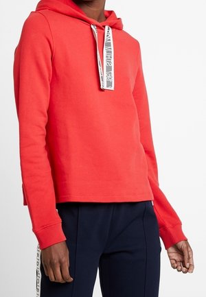 Man draagt een felrode hoodie met witte trekkoorden en zwarte broek met witte zijstrepen, staand tegen een witte achtergrond.