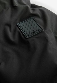 Tissu noir présentant un patch carré texturé en relief avec des bandes diagonales et le texte « 40° 2672° N » embossé dessus.