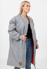 Vrouw in een oversized grijze bomberjack met oranje voering, over een zwart top en blauwe spijkerbroek, staande tegen een witte achtergrond.