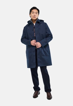 Homme debout portant un manteau à capuche bleu marine, pantalon sombre, chaussures marron, les mains ajustant les boutons du manteau sur un fond uni.