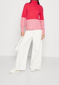 Rosa und korallenfarbener Rollkragenpullover mit geripptem Muster, kombiniert mit weiten weißen Hosen und weißen Turnschuhen, gehalten mit einer kleinen Tasche.