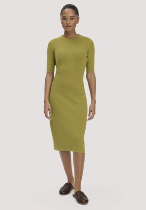 Frau mit dunklen, zurückgebundenen Haaren, die ein figurbetontes, knielanges Strickkleid in Olivgrün und dunkelbraune Slipper trägt, steht und blickt nach vorne.