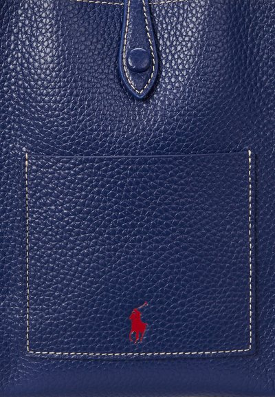 Primo piano di una tasca in pelle blu navy strutturata con un logo rosso di giocatore di polo e una cinghia in pelle cucita con bottone sopra.