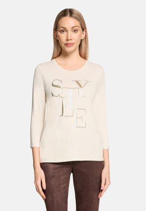 Chemise beige à manches longues avec un design en découpe métallique formant le texte "STYLE". Texture lisse, encolure ronde classique et coupe ajustée.