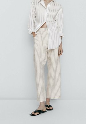 Femme portant un pantalon ample à fines rayures de couleur claire et une chemise oversize rayée assortie, debout avec une main dans la poche et des sandales noires.