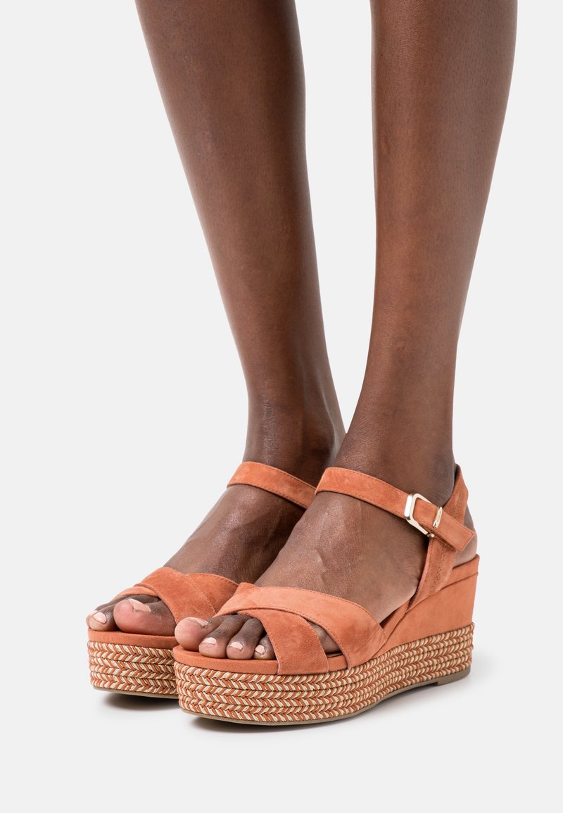 Pieds portant des sandales compensées en daim beige avec des brides à la cheville et des détails tissés sur la semelle sur un fond uni.