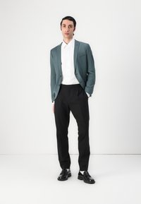 Blazer turchese chiaro abbinato a una camicia bianca, pantaloni neri e mocassini neri. Il tessuto è liscio; il blazer presenta una tasca singola.