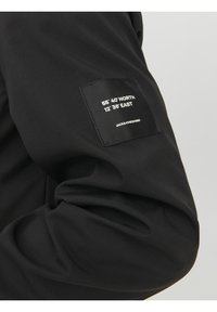 Czarna kurtka o gładkiej fakturze materiału. Na rękawie znajduje się prostokątna naszywka z koordynatami i nazwą marki.